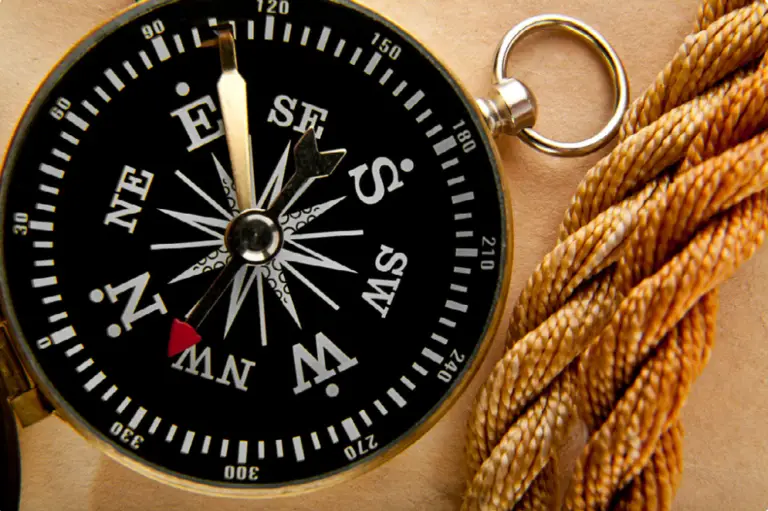 Close-up of a black brass compass with white cardinal marks on a sandy surface, a red needle indicating south, and a coiled rope nearby.